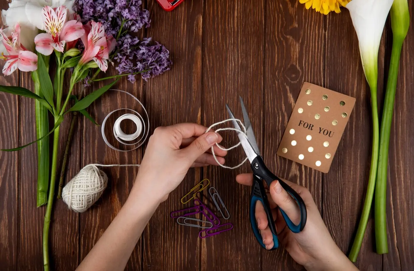 Draufsicht auf Hände, die mit einer Schere eine Schnur schneiden, sowie auf eine Postkarte, Büroklammern und einen Strauß rosa Alstroemerien mit Flieder, alles auf Holzuntergrund.